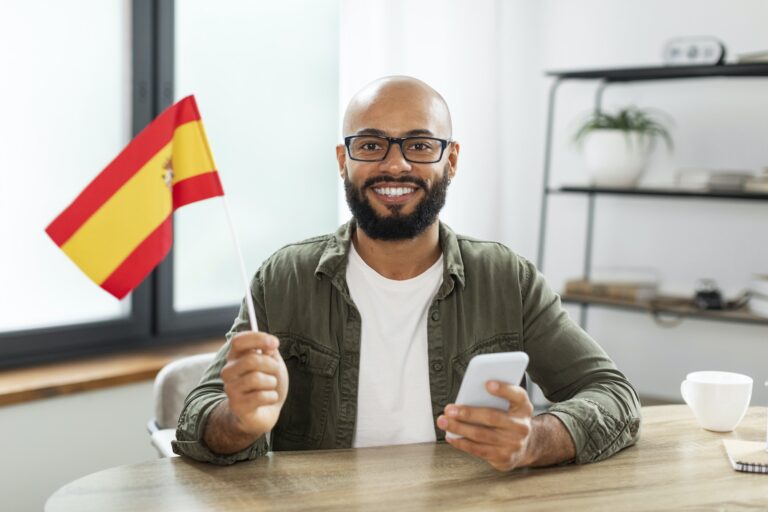 Immigration Services- Excited male tutor sitting at table with flag of Spain and using smartphone, looking and smiling at
